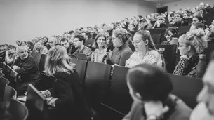 A black-and-white photo of the audience taken from the right side of the lecture hall. A female student is in the process of asking a question.