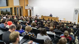 A photo of the lecture hall taken from the upper left part of the room. Parts of the audience are visible as well as David Frum behind the lectern, who is gesticulating with his left hand as he is talking. Also visible is the book table next to the lectern.