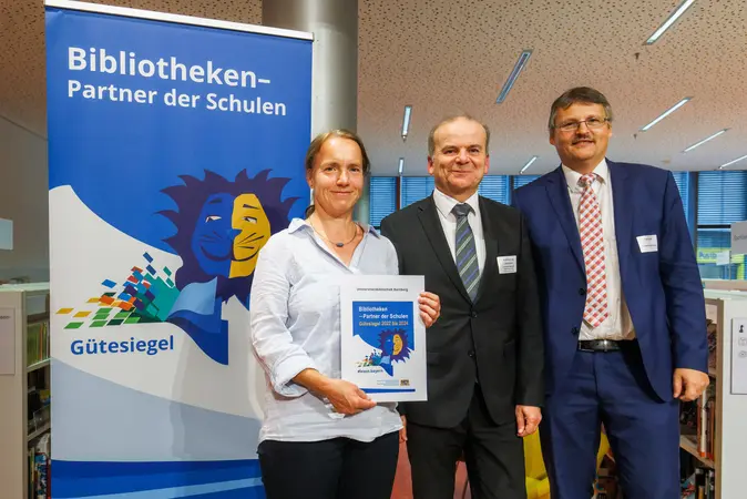 Dr. Fabian Franke (r.) und Christiane Lauterbach von der Universitätsbibliothek Bamberg nehmen das Zertifikat aus den Händen von Ministerialdirektor Dr. Rolf-Dieter Jungk entgegen.
