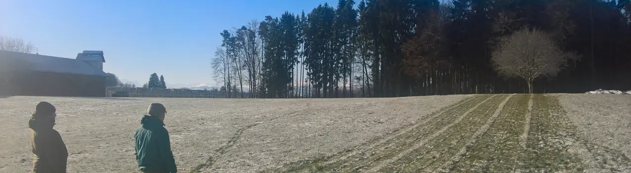 Zwei Studierende stehen auf einem Acker am Waldrand bei schönem Wetter. Es findet eine Geländebegehung statt