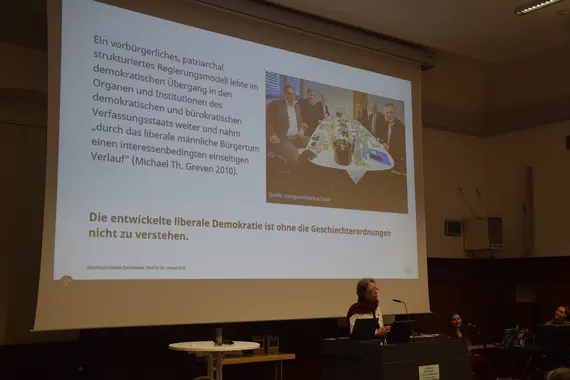 Festrednerin Prof‘in Dr. Ursula Birsl steht vor einem Pult. Hinter ihr ist eine Leinwand. Darauf ist das Zitat zu lesen: "Die entwickelte liberale Demokratie ist ohne die Geschlechterordnungen nicht zu verstehen." 