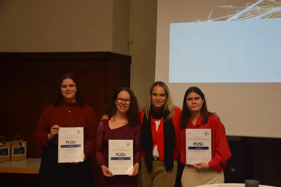 Drei Personen halten eine Urkunde in den Händen. Diese sind die Preisträgerinnen des PUSh-Preis 2025: Judith Albert, Julia Halbritter, Linda Ruppert. Zwischen ihnen steht Silvia Annen, stellvertretende Gleichstellungsbeauftragte in der Wissenschaft 