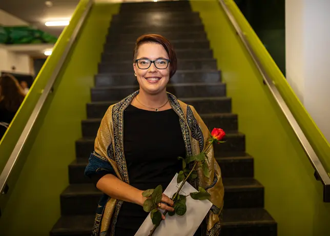 Ein Foto von Dr. Mareike Spychala, eine weiße Frau mit kurzen Haaren, Sidecut und Brille. Sie trägt ein schwarzes Kleid und steht vor einer Treppe.