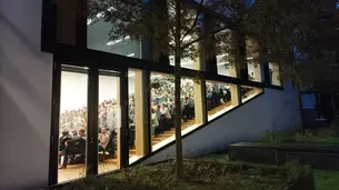 A photo showing the lecture hall MG1/00.04 from outside. One can see the lit-up room and the audience through the large windows.