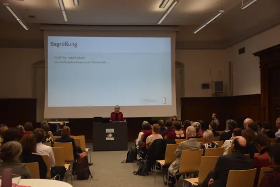 Die Universitätsgleichstellungsbeauftragte in der Wissenschaft, Prof‘in Dr. Astrid Schütz steht vor einem Pult. Hinter ihr ist eine Leinwand, auf der das Wort "Begrüßung" steht. Links und rechts sind Stühle aufgereiht, auf denen die Anwesenden sitzen