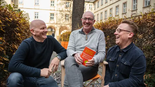 Thomas Weißer, Stefan Huber und Konstantin Lindner mit dem reisenden Regiestuhl auf dem Campus der Universität Bamberg.