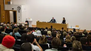 A photo of the lecture hall taken about half-way up the steps. Parts of the audience is visible from behind. David Frum and Prof. Dr. Christine Gerhardt are standing behind the lectern.