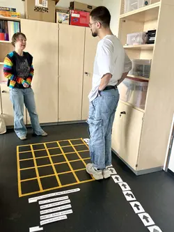 Sanne Grabisch führt mit dem auf einem Feld im Bodenraster stehenden menschlichen Roboter einen Bewegungsalgorithmus aus