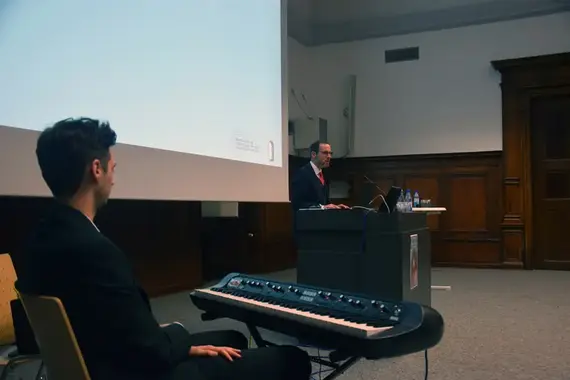 Im Vordergrund dieses Bildes sieht man einen jungen Mann von hinten, der vor einem elektrischen Piano sitzt. Hinter ihm steht ein Mann, der Universitätsprofessor Dr. Kai Fischbach, der vor einem Rednerpult steht und eine Rede hält.Piano 