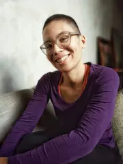 Portrait der Autorin Chantal Fleur-Sandjon, einer Schwarzen Frau mit kurzen Haaren und großer Brille. Sie trägt einen lilanen Pullover und lächelt in die Kamera.