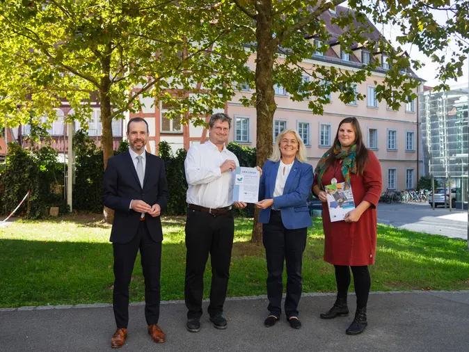 Universitätspräsident Kai Fischbach, Frank Lechner, Leiter des Referats Umwelt/Energie der IHK für Oberfranken Bayreuth, Universitätskanzlerin Dagmar Steuer-Flieser, nd Carolin Brix-Asala, Umweltmanagementbeauftragte der Universität, stehen vor einem Baum im Burgershof. 