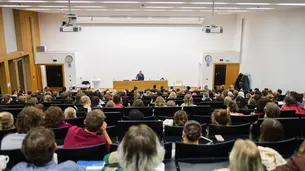 A photo of the lecture hall from the upper end of the room. The audience is visible from behind. David Frum is standing behind the lectern facing the camera.