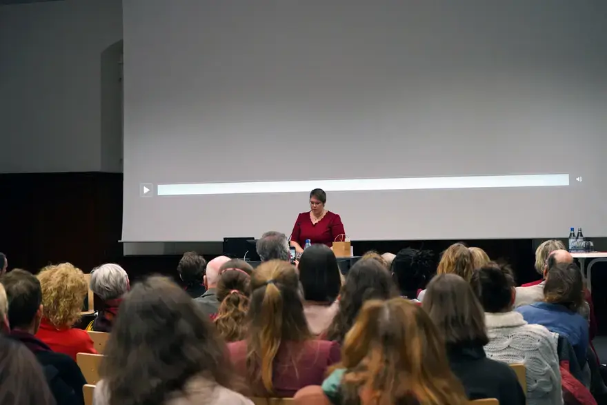 Das Foto zeigt eine Frau, Dr. Kerstin-Anja Münderlein, die vor einem Publikum steht und eine Rede hält. Im Vodergrund sind die Köpfe des Publikums von hinten zu sehen, vor ihnen steht Frau Münderlein an einem Rednerpult. Hinter ihr ist eine große Leinwand.