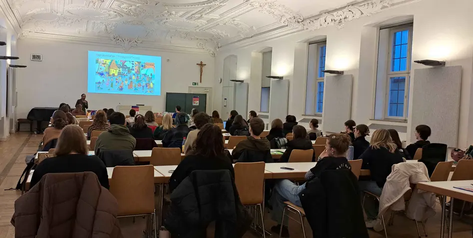 Das Bild zeigt den Hörsaal U5/00.26 von der Eingangstür aus. Man sieht die zahlreichen Zuschauenden der Veranstaltung von hinten und die Sprecherin und ihre Powerpoint (mit einem Bild aus einem ihrer Bücher) vorne.