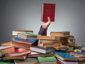 Das Bild zeigt einen Haufen aus Büchern. Oben ist eine ausgestrecke Hand mit einem Buch. Der restliche Körper ist vom Bücherhaufen verdeckt.