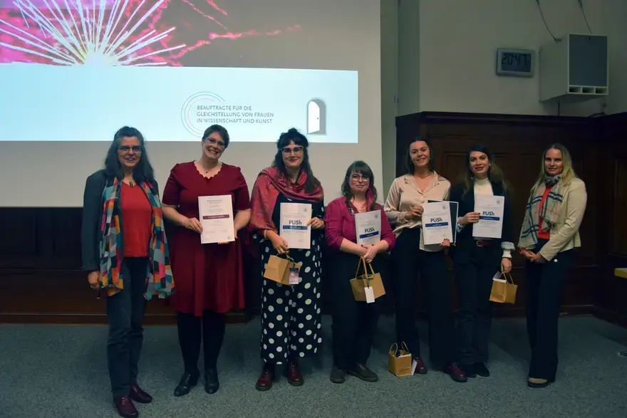 Auf dem Foto sind sieben Frauen zu sehen, die in einer Reihe stehen und lächeln. Die fünf Frauen in der Mitte halten eine Urkunde. Hinter ihnen ist eine Leinwand, die das Logo des Gleichstellungsbüros und der Gleichstellungsbeauftragten für Frauen in Wissenschaft und Kunst zeigt.