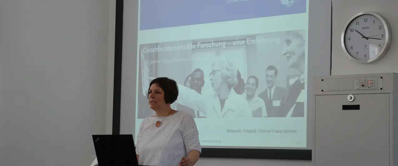 A woman in a white shirt standing behind a table and a laptop. Behind her is a screen projector with the inscription 'Geschlechtersensible Forschung - Eine Einführung'.