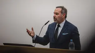 A photo of David Frum, a white man with salt and pepper hair and beard. He wears a suit and tie and is standing behind a lectern while speaking and gesturing with his right hand.