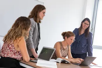 Vier professionell gekleidete Frauen arbeiten gemeinsam an einem Tisch, auf dem aufgeklappte Laptops stehen. Im Hintergrund ist ein Whiteboard zu sehen.