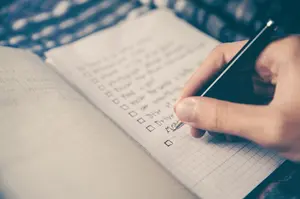 Über einen Block hält eine Hand einen Stift. Auf dem Papier sind bereits mehrere To-Dos abgehakt.