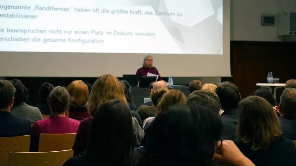 Dieses Bild zeigt eine Frau, Dr. Gisela Mettele, die einen Vortrag hält. Im Vordergrund sind die Köpfe des Publikums zu sehen, hinter ihnen steht Frau Mettele vor einer Leinwand