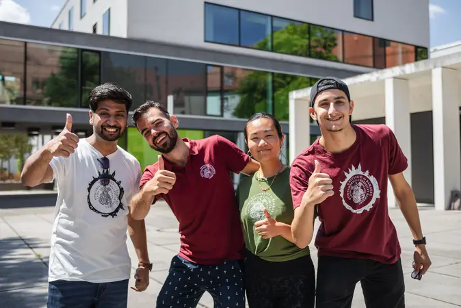 Studierende zeigen vor einem Universitätsgebäude den Daumen nach oben.