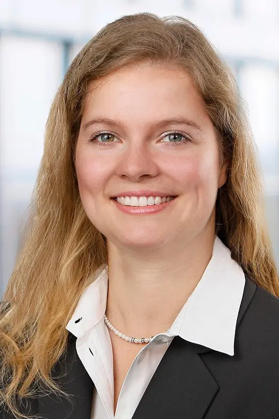 Eine lächelnde dunkelblonde Frau mit schulterlangen Haaren, weißer Bluse, schmaler Perlenkette und schwarzem Blazer
