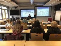 Studierende sitzen in einem Seminarraum mit dem Rücken zur Kamera. Vor der interaktiven Tafel stehen Leonie Ackermann und Hannah Deininger. 