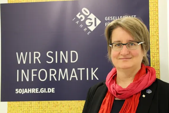 A blond women with red scarf in front of a poster with the text "Wir sind Informatik" (50jahre.gi.de)
