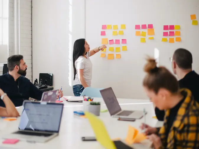 Frau präsentiert Seminarteilnehmer*innen bunte Post-Its auf einer weißen Wand. 