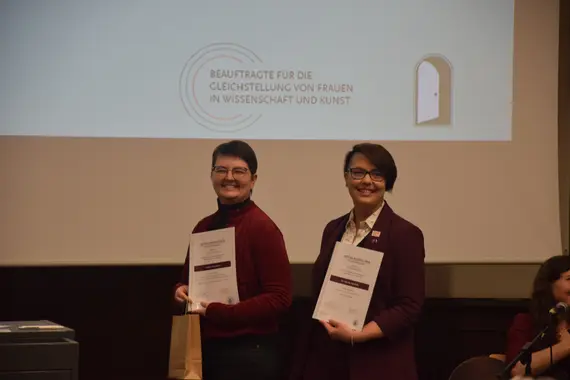Zwei Personen stehen vor einer Leinwand, lächeln und halten eine Urkunde auf dem Arm. Diese sind die Preisträger*innen des Bettina-Paetzold-Preis: Leonie Ackermann und Dr. Mareike Spychala