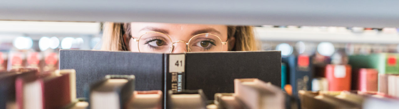 Studentin in der Uni-Bibliothek schaut in ein aufgeschlagenes Buch zwischen Bücherregalen
