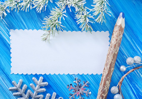 White card on a wooden, blue background. Fir bough upper edge of the frame, silver stars and bough with frosted berries at the lower edge. On the right of the card is a wooden pencil with a blue tip.